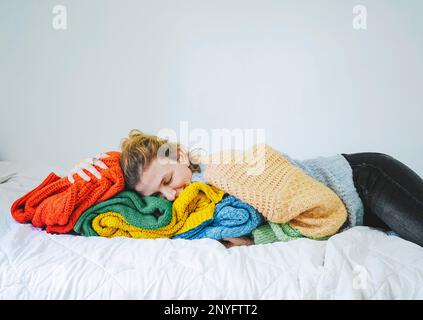 Calm young female in warm clothes lying on colorful knitted sweaters on soft bed with closed eyes at home Stock Photo