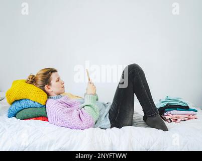 Full body side view of positive young female wearing warm colorful knitted sweater and jeans lying in bed resting head on colorful pile of multicolore Stock Photo