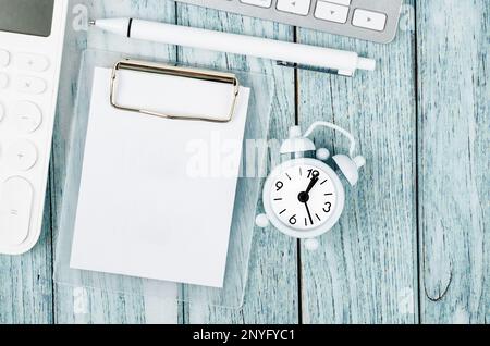 Small white paper blank page on clip board, alarm clock and calculator on blue wooden background using as meeting note, writing message or task list o Stock Photo