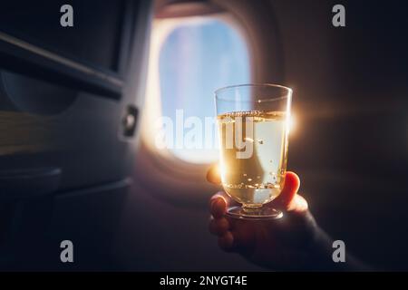Man enjoying drink during flight. Passenger holding glass of sparkling wine against airplane window. Stock Photo