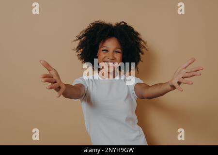 Nice to meet you. Overjoyed happy african woman 20s standing isolated on beige background with hands wide open, kind positive female in white tshirt w Stock Photo