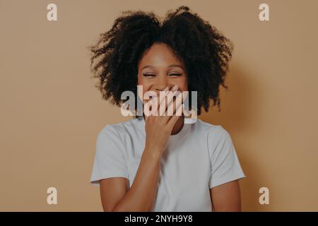 Close up studio portrait of happy biracial girl. Beautiful dark skinned millennial woman covering her mouth with hand while laughing. Cute positive mi Stock Photo
