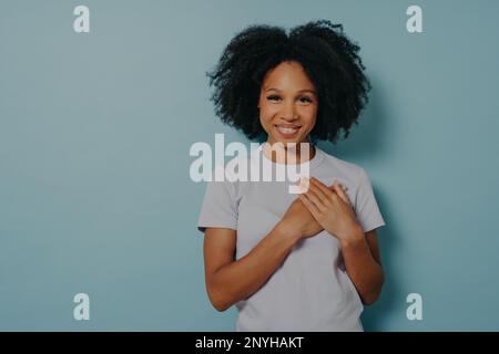 Grateful hopeful happy black woman holding hands on chest, satisfied and sincere african lady expressing love from heart, isolated on blue studio back Stock Photo