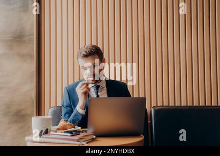 Male office manager wearing blue suit and white shirt, speaking to his workers in online meeting and looking at laptop computer while sitting and work Stock Photo