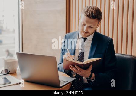 Concentrated entrepreneur in formal outfit wearing earphones watching online webinar on laptop taking notes of important information, writing everythi Stock Photo