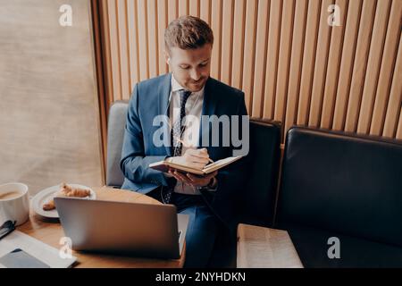 Young bearded businessman in formal wear sitting in cafe, writing down notes in notebook while listening online lecture for educational seminar on lap Stock Photo