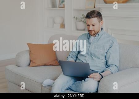 Attentive handsome young man in casual clothes using laptop during leisure time while sitting on sofa at home, looking for his favorite TV shows onlin Stock Photo