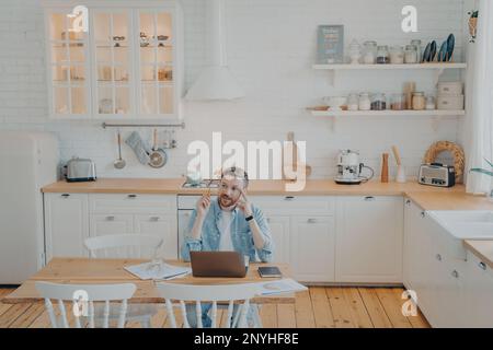 Young office worker working remotely from home with computer, sitting by kitchen table, tasked with important project, trying to find out solution, wr Stock Photo