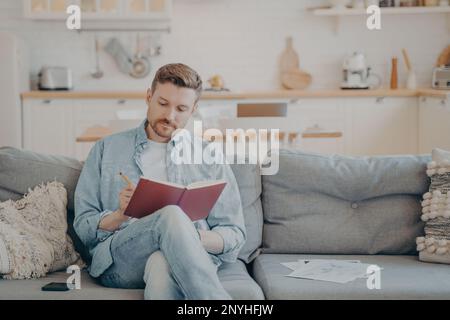 Young man in casual clothes sitting on couch with legs crossed, writing down information in red note book, taking important notes with pencil, organiz Stock Photo