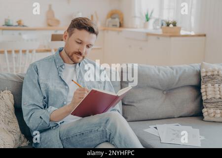 Busy young man dressed in casual clothes writing in red notebook with pencil while sitting on comfortable couch with crossed legs, looking focused, ki Stock Photo