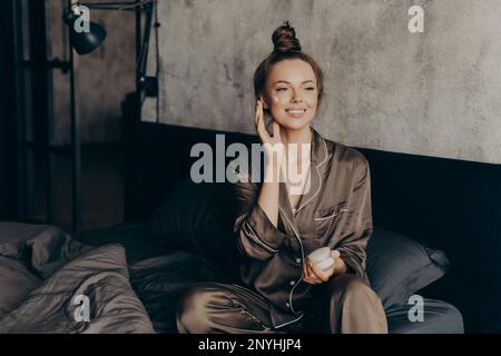Young pretty european woman in silk pajama takes care of skin and body after waking up in morning at home while applying moisturizing face cream, gett