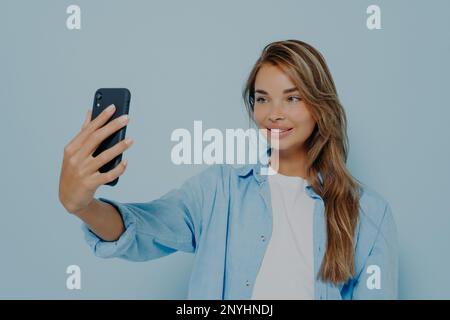 Time for selfie. Magnificent young woman in stylish casual clothes with long dyed hair taking photo of herself on modern cell phone, looking charming Stock Photo