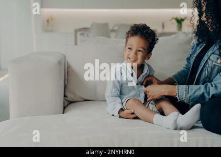 Young caring mother dressing up her little cute son with curly hair, putting on adorable boy shirt and buttoning it, getting ready to walk in park whi Stock Photo