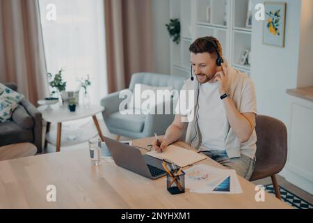 Happy young man adjusting headset while studying online on laptop or having web conference during remote work at home, male freelancer having video ca Stock Photo