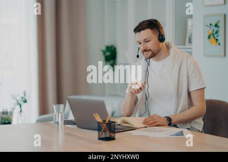 European male student learns foreign languages online has video lesson uses modern headset focused at laptop screen computer engaged in conference lis Stock Photo
