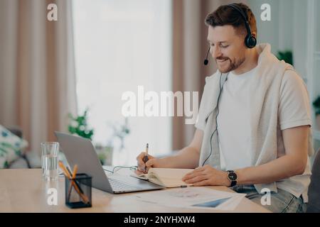 Side view of smiling man in headset having video lesson online, writing some notes in notebook during elearning at home. People and distant education Stock Photo