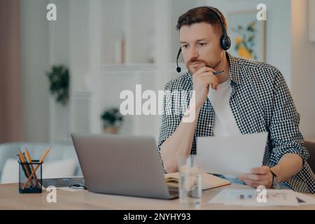 Serious man freelancer in casual wear holding document in hand and looking at laptop screen, analyzing information while working online in living room Stock Photo