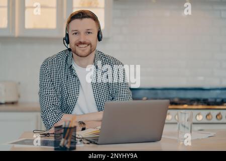 Handsome glad man customer support manager gives advice or consultation to clients works online from home poses at home kitchen wears wireless headpho Stock Photo