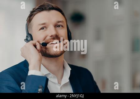 Close up of happy office worker handsome male call center operator wearing headset and working in office, talking with client online with positive fac Stock Photo