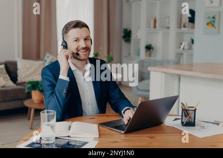 Positive bearded young man freelancer wearing suit in headphones with microphone smiling at camera while sitting at his workplace at home, using lapto Stock Photo