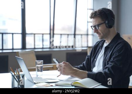 Young german male freelancer in headphones and glasses looking at smartphone display in hands, checking last emails or typing message sms while sittin Stock Photo