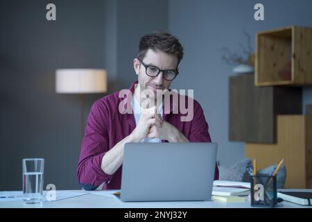 Pensive young German man home office employee sits at desk at workplace operates online on laptop pondering thinking. Focused positive male looks at c Stock Photo