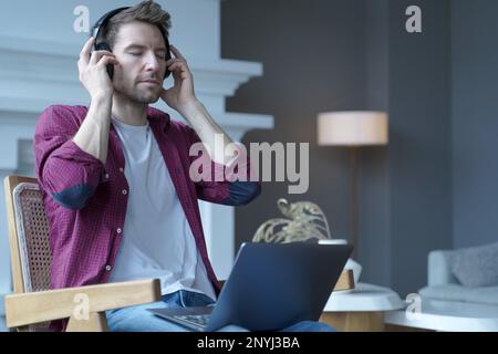 Young austrian guy sitting on wooden wicker chair with open laptop on his lap listening music in wireless headphones totally immersed in favorite, sou Stock Photo