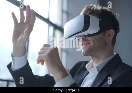 European man office employee wears formal suit and VR headset glasses excited of experiencing virtual reality stretching his hands up trying to catch Stock Photo