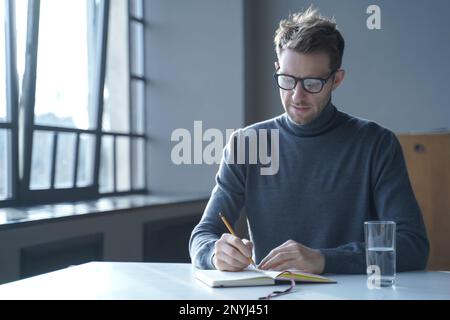 Confident focused young German man freelancer in glasses taking notes in agenda while sitting at desk at home interior, has break from work online, wr Stock Photo