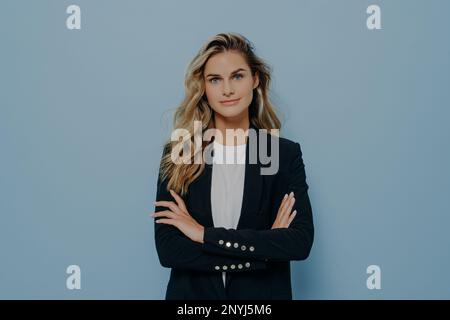 Young confident blonde business woman with long wavy hair dressed in smart black costume standing with crossed arms, posing over blue background in st Stock Photo