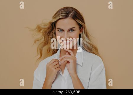 Close up portrait of flirty romantic and feminine beautiful blond woman looking at camera playfully and with bright wide smile, holding hands in front Stock Photo
