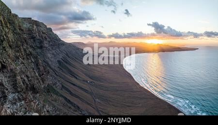 Aerial panorama of sunrise on the coast of Famara, a holiday destination in Canary Islands, Spain, captured from rocky slopes with clouds n calm water Stock Photo