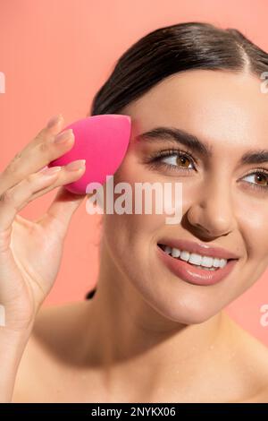 cheerful young woman applying makeup foundation with beauty sponge isolated on pink,stock image Stock Photo