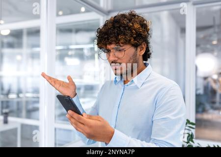 Close up of businessman in middle of office holding phone, man upset reading bad news online, hispanic man disappointed and depressed by notification. Stock Photo