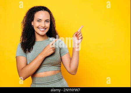 Happy mixed race sporty curly woman, dressed in sports outfit, pointing fingers of both hands to side at empty space for your presentation, looks at camera, smile, stands on isolated yellow background Stock Photo