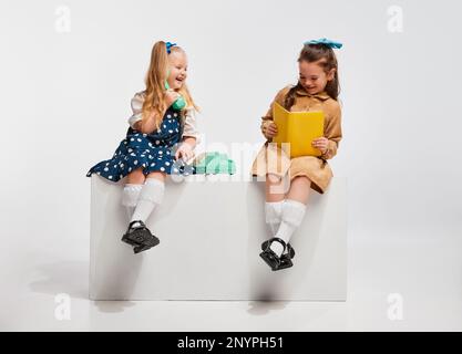 Playful happy smiling little girls talking on phone and reading book against grey studio background. Concept of childhood Stock Photo