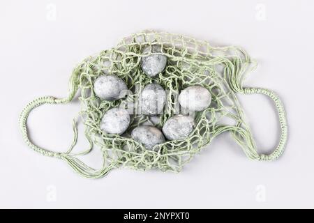 Grey Easter eggs in a green string bag on a grey background. Minimalism, copy space, flat lay Stock Photo