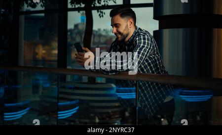 Young man businessman caucasian hispanic male client guy stand in modern business office hold smartphone chatting online use mobile phone banking app Stock Photo