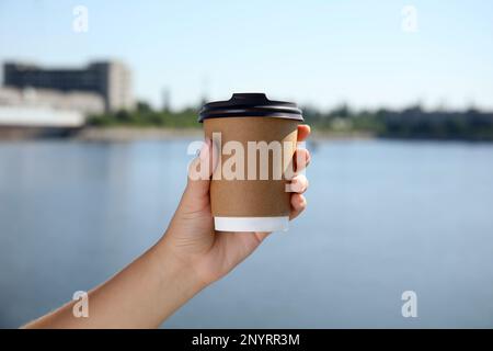 Woman holding takeaway cardboard coffee cup with plastic lid against blue river, closeup Stock Photo