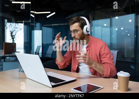 Worried young male freelancer, programmer in headphones, sitting at a table and working on a laptop, talking on a video call. Problems in the office. Stock Photo