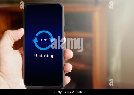 Male hand holding mobile phone showing update interface and icon with percentage progress on the screen. Man updating smartphone at home. Stock Photo