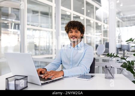 Portrait of young programmer inside office with laptop, indian man smiling and looking at camera, man typing on keyboard, writing code for software, happy with achievement results. Stock Photo