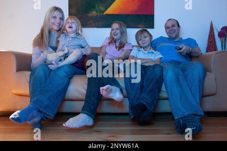 Family Life, TV Comedy. Fun and laughter on the faces of a young family sitting watching television at home. TV POV. From a series of related images. Stock Photo