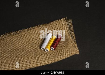 Still life with spools of thread of three colors on a folded burlap cloth on a black surface Stock Photo