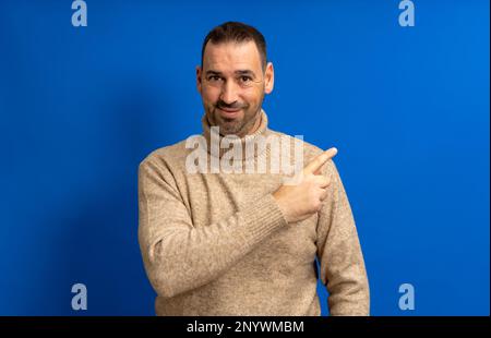 Friendly bearded Hispanic man in his 40s wearing a beige turtleneck pointing to the side, isolated over a blue background Stock Photo