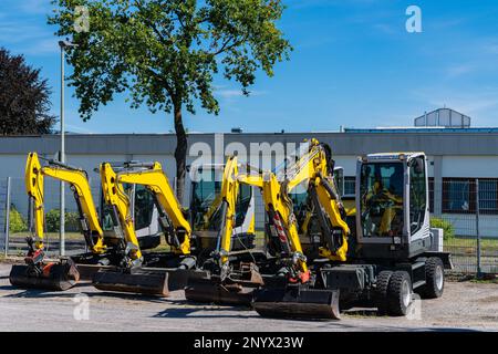 Small excavators standing in a row. Building under the blue sky in the background. Stock Photo