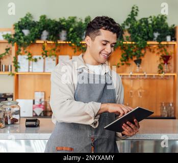 Smiling barista in an apron holding a digital tablet while leaning counter. Coffee shop owner typing on digital tablet. Stock Photo