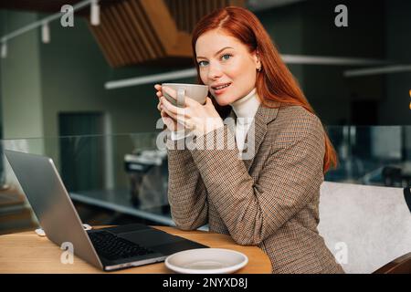 Young woman with ginger hair holding a cup. Female entrepreneur drinks coffee while working from a coffee shop. Stock Photo