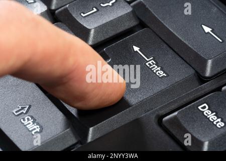 Finger pressing the enter key on a simple black computer keyboard, object macro, extreme closeup, side view. Confirmation, sending a message, chatting Stock Photo
