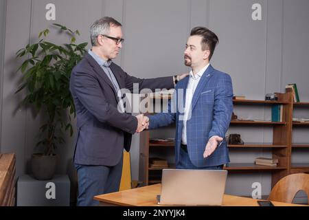 Two happy business partners in blue suits smiling and making handshake in office. Businessman pointing on the laptop Stock Photo